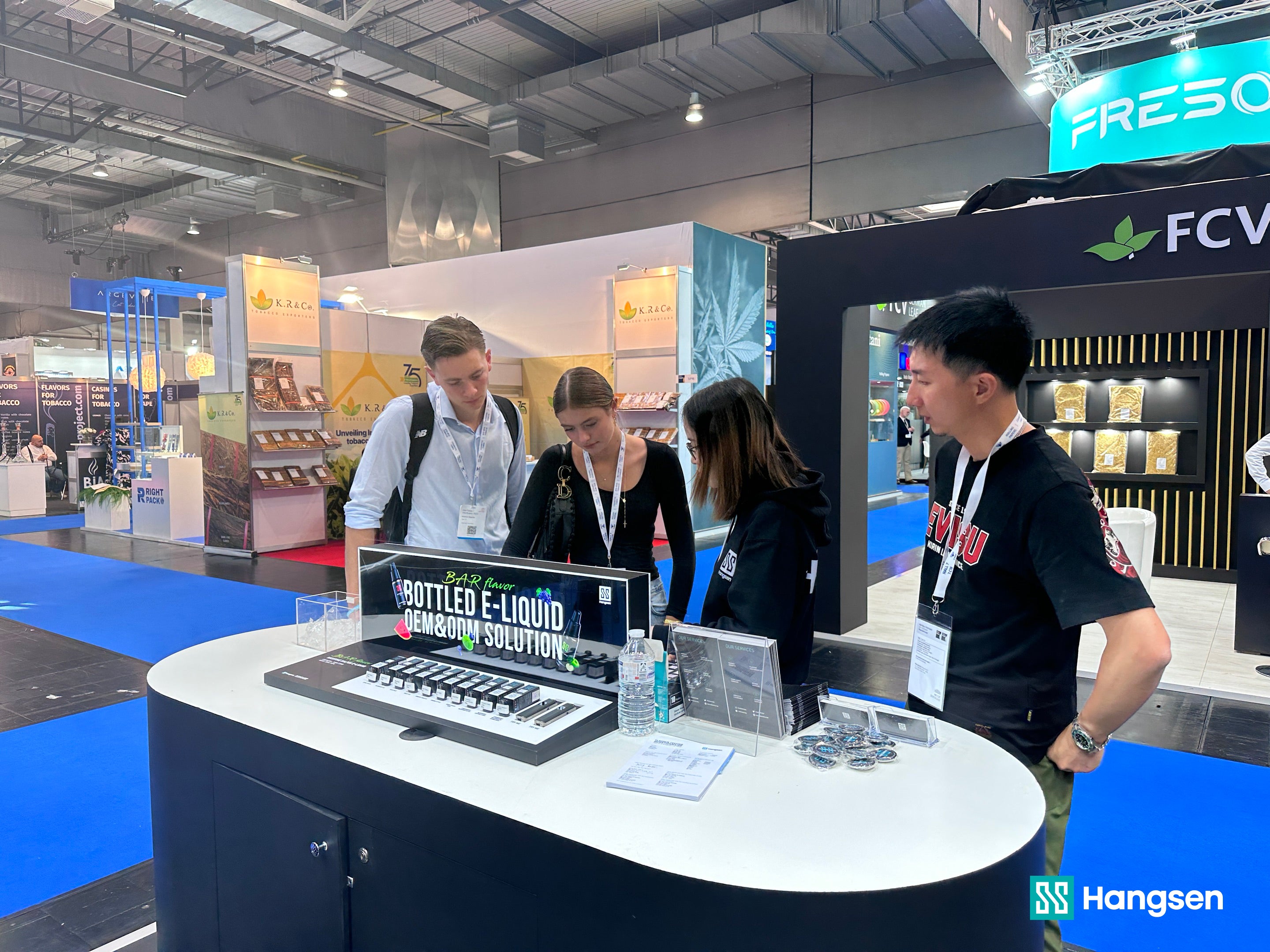 Attendees explore Hangsen's Bar Flavor Bottled E-Liquid OEM&ODM Solution at a trade show booth, with a sign prominently displayed in the foreground. The booth is staffed by two individuals engaging with visitors amidst a bustling exhibition hall.