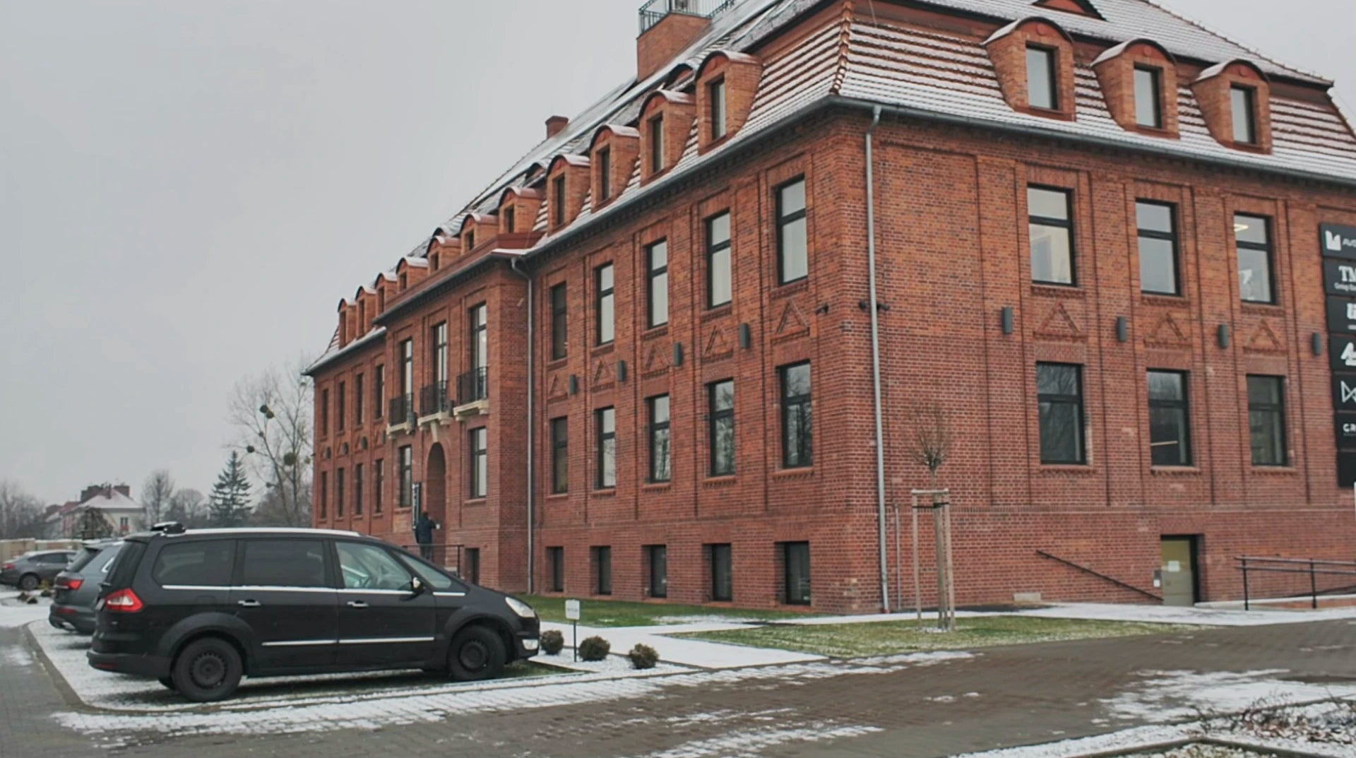 A red brick factory building with a sloped roof and multiple dormer windows, located in a snowy setting. There is a parking lot in front with a few cars, most prominently a black minivan. The sky is overcast, suggesting a cold day.