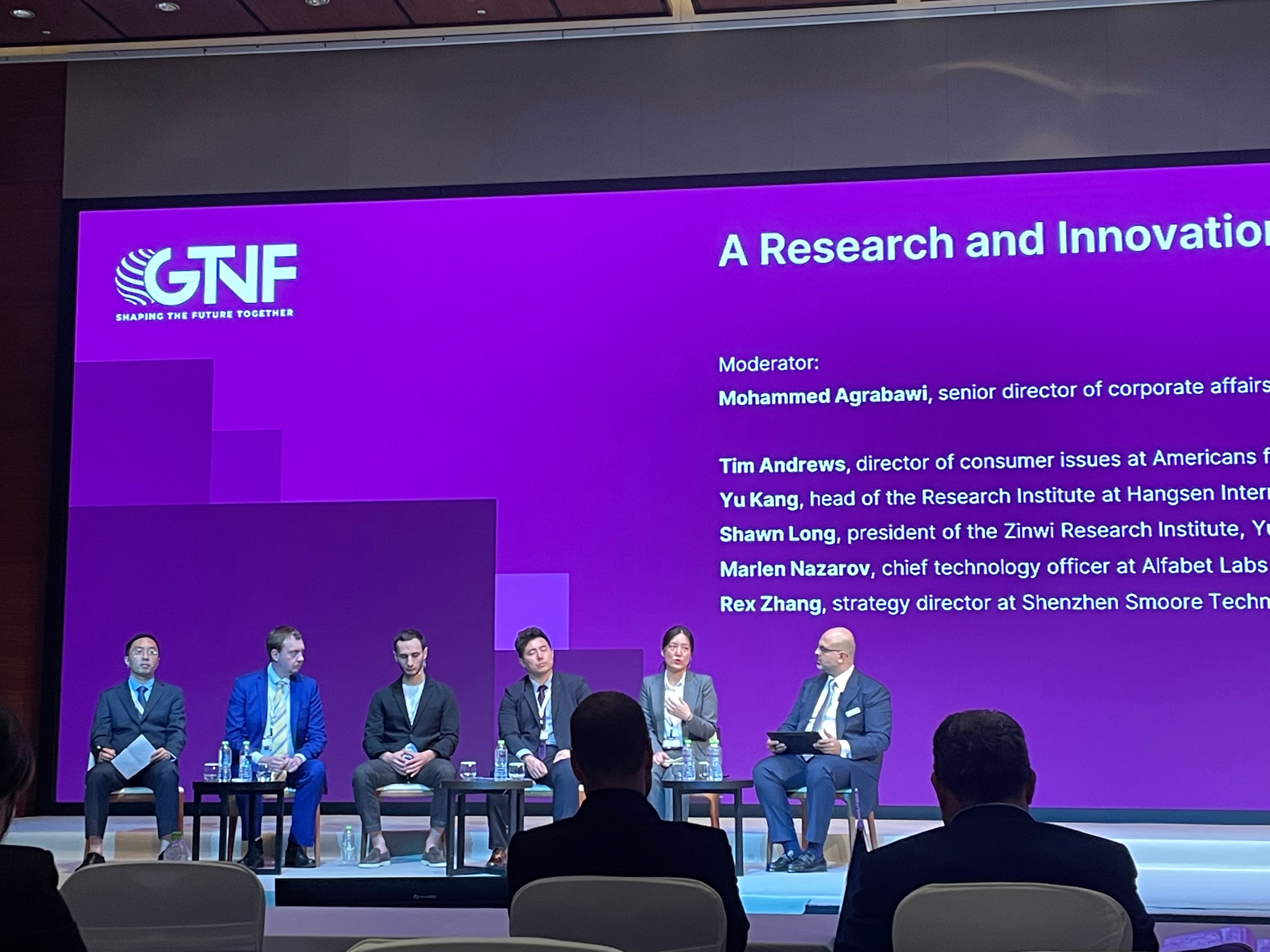 Panelists are seated on stage at the GTNF conference during a session on research and innovation, with the session's title and the names of the moderator and speakers displayed on the screen behind them.